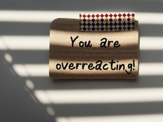 Note stick on wall with handwriting YOU ARE OVERREACTING, concept of gaslighting, person who presents false narrative to leads another to doubt their perceptions and become distressed and dependent  