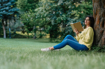 Naklejka na ściany i meble Young woman reading book near tree in park