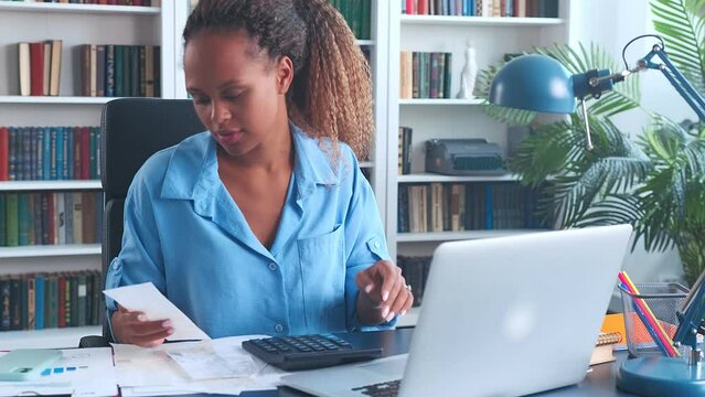 Young carried away African American woman entrepreneur prepares tax reports using calculator and laptop to record data from cashier checks and expense documents sits at desk in home office
