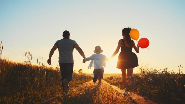 Rear view, happy family runs together on wheat field holding hands. Mom holding colour balloons. Summer adventure, journey, vacation in countryside outside city. Enjoy rest, freedom. Walk at summer.