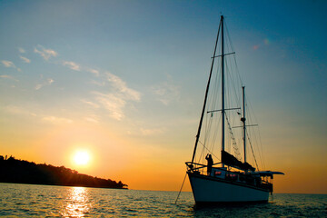 A yacht is silhouetted by the rising sun as it is anchored near an island and a man stands on deck.