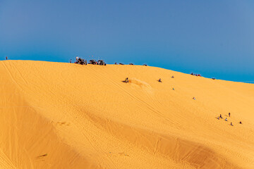 Fototapeta na wymiar Unrecognizable far people sitting and sunbathing, a man is riding a quad in the gold colored sand dune with buggy cars parked in the summit. Sahara desert of Taghit, Algeria. Sunny day with a blue sky