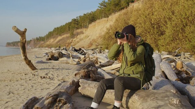 Pretty girl sits on log on river bank, takes photos. Young lady portrait in beanie, jacket sitting on sandy beach near dry log seeing at pictures on screen