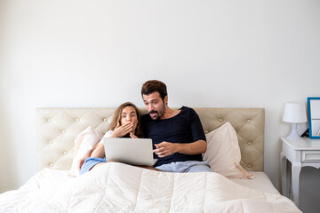 Fototapeta premium Happy Couple watching movie by laptop. Young couple watching a movie on bed at home in the afternoon. Man and woman watching movie from computer notebook on their bed.