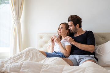 Couple relaxing on bed at home. Lover cuddling and embracing while sitting in bed in the morning. Romantic couple sitting on bed in bedroom. Young married Couple relaxing in bed with hot drink.