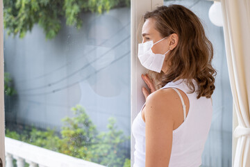 Woman wearing protective mask feeling sad sitting on bed at home. Female wearing a protective mask...