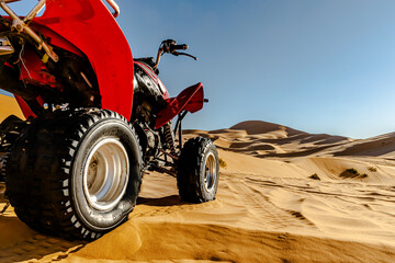 Quad bike orange color vehicle parked in the Sahara desert, selective focus in foreground, golden yellow colored sand dunes and blue sky in blur background. Ground level rear view of the side part.