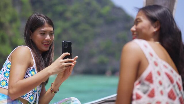 Asian woman friend using mobile phone taking picture together during travel on boat passing island beach lagoon in sunny day. Attractive girl enjoy and fun outdoor lifestyle on summer holiday vacation
