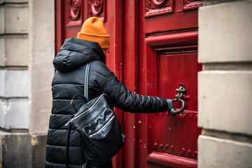 A beautiful girl in a black coat and orange hat walking through the streets of the city of Paris found a beautiful old red door. Door with antique brass handle. The girl is trying to open an old door 