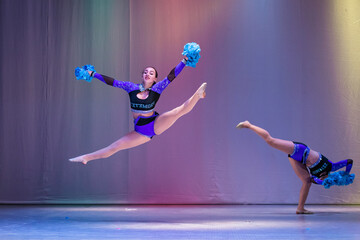 athletes perform on stage, young cheerleaders perform at the cheerleading championship, girls in a jump, girls are holding pompons, hands raised up, toe touch, girl doing acrobatic and flexible tricks