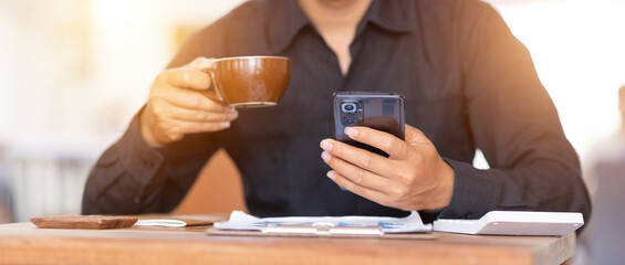 Young Asian man in black long-sleeved shirt sitting working in a cafe Use your smartphone and documents on the table.