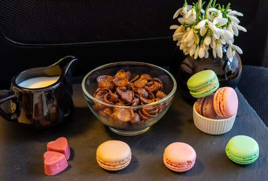 Breakfast with chocolate cereals, milk, macaroons,  pink heart shaped chocolate pralines and snowdrops in a black vase, on a black background