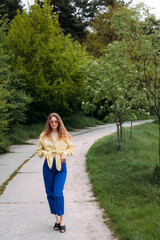 A girl with blond curly hair dressed in blue jeans and a yellow shirt, wearing sunglasses, stands on a path in a picturesque park in summer.