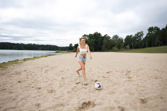 A beautiful slender girl with long blond hair and long legs runs along the beach towards a soccer ball, wearing short sexy denim shorts and a white tank top.
