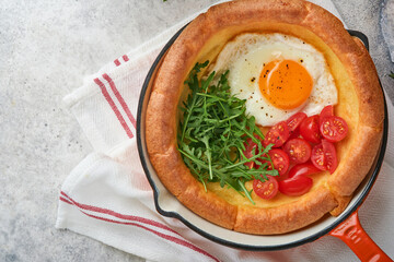 Dutch baby pancake. Fresh homemade Dutch Baby pancake with fried egg, tomato and green arugula in reed cast-iron pan on light gray concrete rustic table. Flat lay, close up.