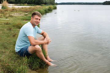 A smiling man with an athletic body, dressed in denim shorts and a blue t-shirt, sits on the banks of a picturesque river during the day.