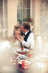 Art work. Beautiful smiling newlyweds in stylish outfits are sitting at the wedding table set against the backdrop of the beautiful interior of the restaurant.