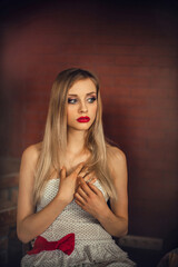 Fototapeta premium A chic young slender girl with blond long hair and bright makeup sits on a chair in the room, against the background of a burgundy brick wall.