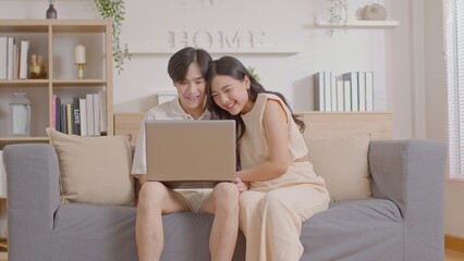 Asian young couple sit on couch or sofa looking at Computer Laptop smile and laughing together. Cheerful lover surfing internet and social media spending time in holiday at cozy home. Valentine Day