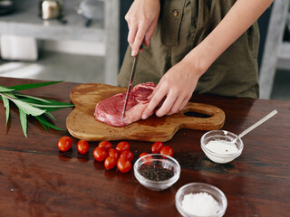 Woman with knife in hand cutting steak meat for frying in kitchen with salt pepper and other spices on table, red cherry tomatoes and herbs, dinner preparation.