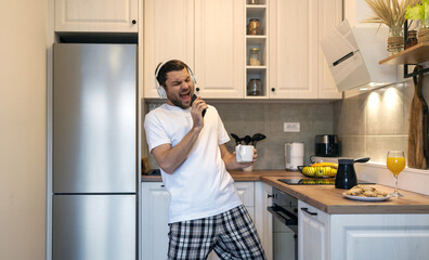 Young man in headphones singing in kitchen in morning using his mobile phone as a microphone.