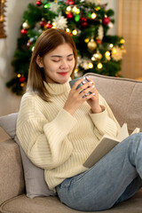 young asian woman holding a cup of coffee and a book on the cozy sofa