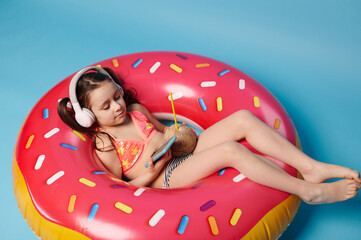 Happy little girl in swimsuit and headphones, relaxing on swim donut ring, drinking coconut water...
