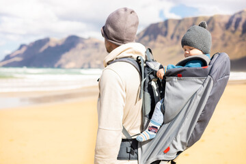 Fototapeta na wymiar Young father carrying hisinf ant baby boy son in backpack on windy sandy Famara beach, Lanzarote island, Spain. Family travel and winter vacation concept