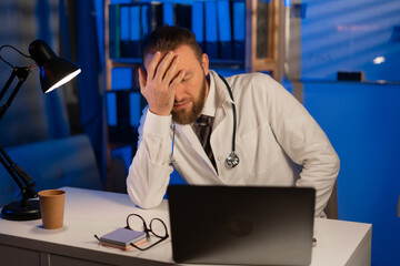 Male tired doctor with migraine overworked, overstressed working at night. sad unhappy health care professional with headache stressed sleepy - obrazy, fototapety, plakaty