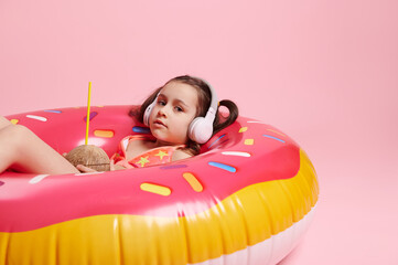 Relaxed little girl with wireless headphones and coco water on pink inflatable swim tube, isolated...