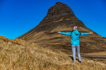 Naklejka premium Girl in blue jacket with outstretched arms admiring migthy stunning iconic Kirkjufell (hat) mountain. Western fjords, Iceland, Europe