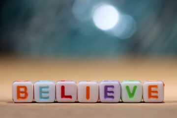 Square beads spelling the word believe against a bokeh blue grey background with light points. Business motivation and inspiration with copy space above.