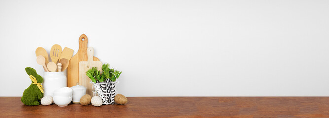 Easter theme kitchenware, utensils and decor on a wood shelf or counter. Banner against a white wall background with copy space. Spring home kitchen cooking concept.
