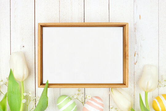 Rustic wood frame with pastel Easter eggs and white spring tulip flowers. Overhead view against a white wood background. Copy space.