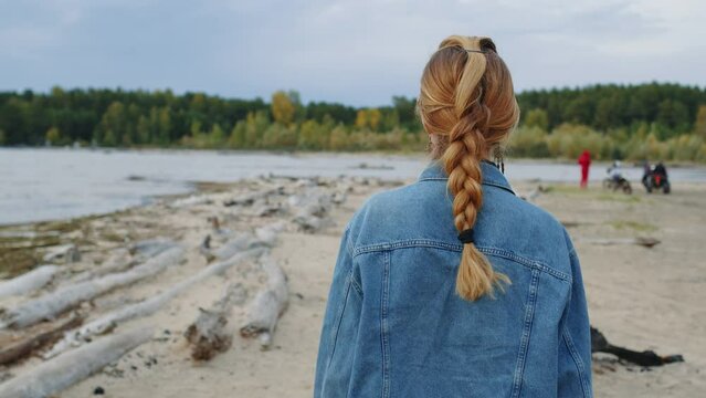 Beautiful girl walking along sandy river bank in cloudy weather. Pretty young woman going on seashore, relaxing, enjoying views of nature. Blonde in jeans jacket rests in promenade. Back view