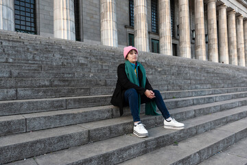 Fototapeta premium Female latin student with brown hair and pink hat sitting on the stairs in college building