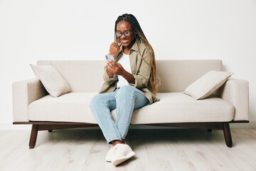 Fototapeta na wymiar African American woman business freelancer working sitting on the couch at home in the phone, business calls and messages happiness smile, home clothes and eyeglasses, light interior background.
