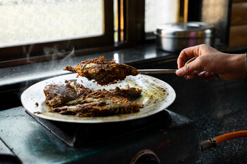Indian Mackerel Fish marinated with spices frying on metallic on aluminium utensil and one piece of fish lifted with help of spoon with woman hand in frame. Used selective focus.