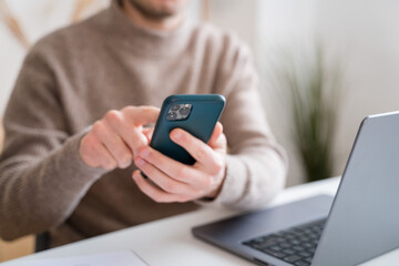 Crop man using smartphone while working on laptop at home