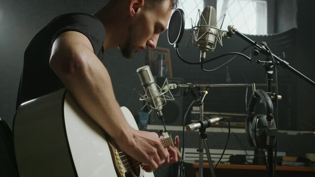 Guitarist sits next to professional microphone, plays guitar. Male singer in recording studio. Vocalist working on music album in sound department. Bearded caucasian man performs song