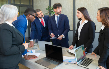 Successful entrepreneur focusing on work with charts during meeting with multiethnic employee. Mature businessman discussing work at conference table in office with senior businesswoman.