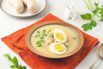 Polish Easter soup Zurek with white sausages and a boiled egg in a green ceramic bowl on a light concrete background. Traditional Polish Easter dishes.