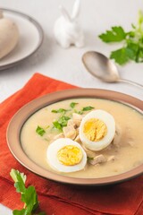 Polish Easter soup Zurek with white sausages and a boiled egg in a green ceramic bowl on a light concrete background. Traditional Polish Easter dishes.