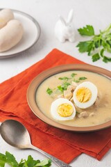 Polish Easter soup Zurek with white sausages and a boiled egg in a green ceramic bowl on a light concrete background. Traditional Polish Easter dishes.