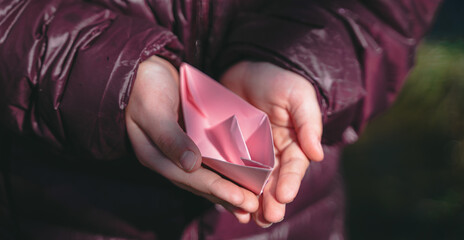 Girl in purple jacket playing with paper boat. Colorful pink ship in hands. Children play, have fun outdoors in spring snow puddle on street. Warm wet rainy weather, old grass. Hello spring, autumn