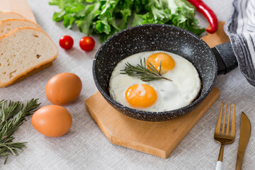 Fried eggs in a frying pan with rosemary, bread, eggs and green salad on a linen tablecloth. Breakfast, lunch and dinner. The view is soft, selective focus. Rustic style.