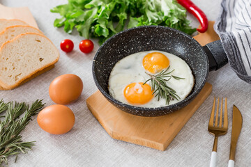 Fried eggs in a frying pan with rosemary, bread, eggs and green salad on a linen tablecloth. Breakfast, lunch and dinner. The view is soft, selective focus. Rustic style.