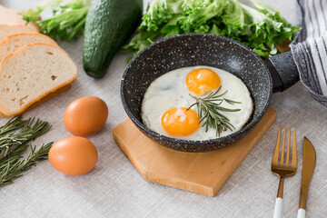 Fried eggs in a frying pan with rosemary, bread, eggs and green salad on a linen tablecloth. Breakfast, lunch and dinner. The view is soft, selective focus. Rustic style.