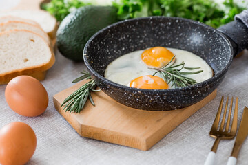 Fried eggs in a frying pan with rosemary, bread, eggs and green salad on a linen tablecloth. Breakfast, lunch and dinner. The view is soft, selective focus. Rustic style.
