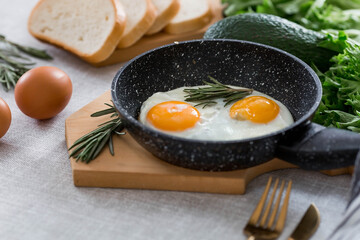Fried eggs in a frying pan with rosemary, bread, eggs and green salad on a linen tablecloth. Breakfast, lunch and dinner. The view is soft, selective focus. Rustic style.